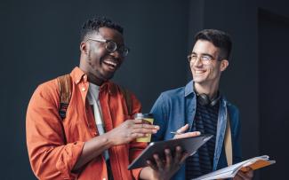 Étudiant souriant tenant une tablette numérique et une tasse de café, debout qui rie avec autre étudiant