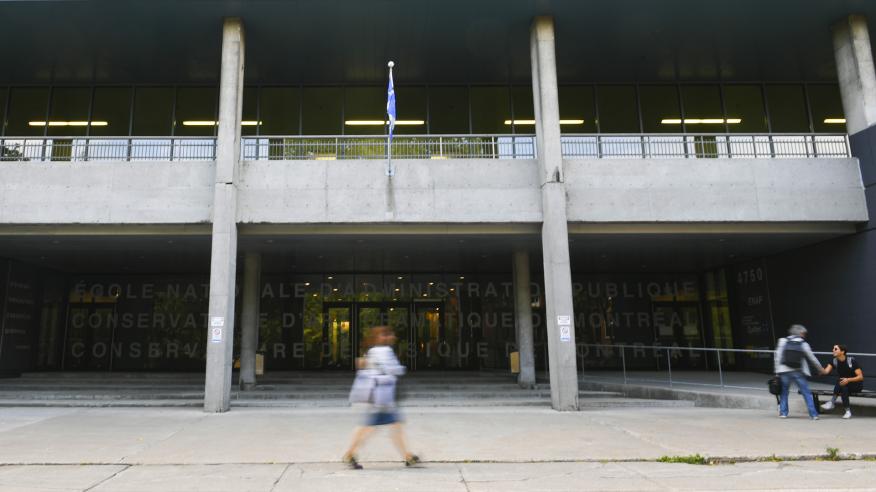 façade du campus de Montréal, femme en floue au centre de la photo et deux personnes à droite se serrant la main